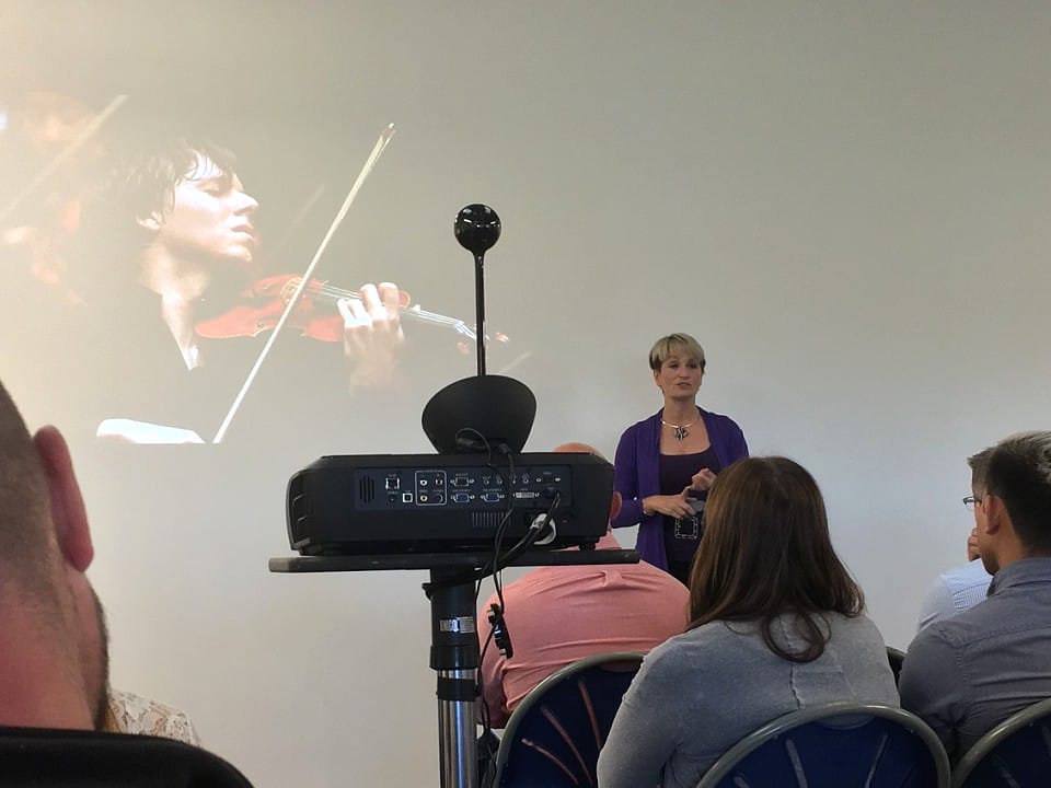 A woman giving a talk with a violinist on the screen